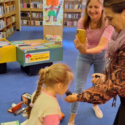 Po prawej stronie stoi bibliotekarka. Trzyma kałamarz. Z lewej strony dwoje dzieci wącha atrament. W tle stoją regały z kolorowymi książkami. Kliknięcie powoduje powiększenie zdjęcia.