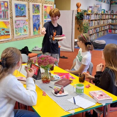 Uczestniczki DKK grają w kalambury. Ruda dziewczynka stoi z książką w ręce i pokazuje palcem. Pozostałe dziewczynki siedzą przy stole. Kliknięcie w obrazek spowoduje powiększenie.