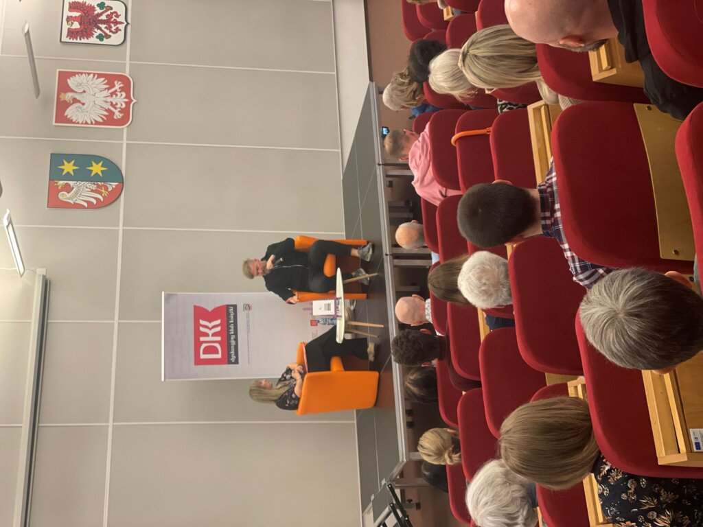 Siedząc na pomarańczowych fotelach na podium rozmawiają Tomasz Organek oraz prowadząca. Na pierwszym planie publiczność zgromadzona w sali.