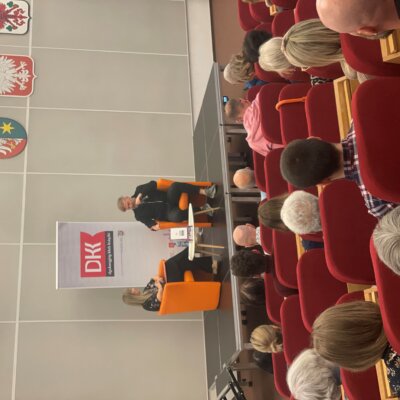 Siedząc na pomarańczowych fotelach na podium rozmawiają Tomasz Organek oraz prowadząca. Na pierwszym planie publiczność zgromadzona w sali.