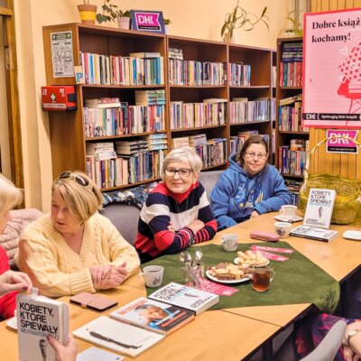 Wnętrze Filii nr 8. Trwa spotkanie Dyskusyjnego Klubu Książki dla dorosłych. Przy stole siedzi siedem kobiet. Na stole leżą książki o Wojciechu Młynarskim. Moderatorka trzyma omawianą książkę – „Kobiety, które śpiewały Młynarskiego" J. Nojszewskiej. Kliknięcie w obrazek spowoduje powiększenie obrazu.