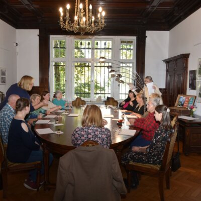 Przygotowanie do warsztatów kaligrafii. Uczestnicy siedzą dookoła owalnego stołu w zabytkowej części biblioteki, nad stołem zapalony żyrandol, w tle duże okno. Kliknięcie powoduje powiększenie zdjęcia.