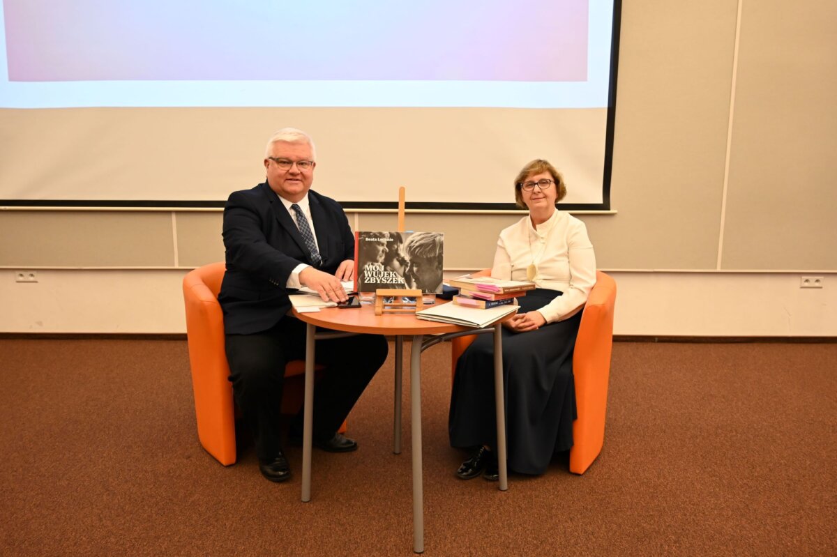 Na zdjęciu widać dwie osoby siedzące przy okrągłym stole. Mężczyzna po lewej stronie ma na sobie ciemny garnitur i białą koszulę, a kobieta po prawej stronie ubrana jest w białą bluzkę i ciemną spódnicę. Oboje siedzą na pomarańczowych fotelach. Na stole znajdują się książki, a jedna z nich jest otwarta i stoi pionowo, z widoczną czarno-białą okładką. W tle widać ekran projekcyjny.