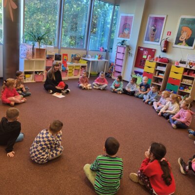 Na zdjęciu znajduje się grupa dzieci siedzących w kręgu, a w jego centrum Pani Bibliotekarka. W tle widać duże okna z widokiem na drzewa. Na ścianach wiszą kolorowe plakaty z postaciami z kreskówek. W pomieszczeniu można zauważyć kolorowe półki z różnymi materiałami. Kliknięcie w obrazek spowoduje powiększenie.