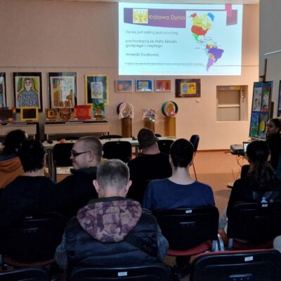 Na zdjęciu widzimy uczestników warsztatów, siedzących na krzesłach, jak również prowadzącą Panią Bibliotekarkę, która omawia prezentację dotyczącą dyni. W pomieszczeniu panuje półmrok, uczestnicy reagują pozytywnie na tematykę prezentacji. Na ścianie widoczna prezentacja multimedialna. Kliknięcie powoduje powiększenie zdjęcia.