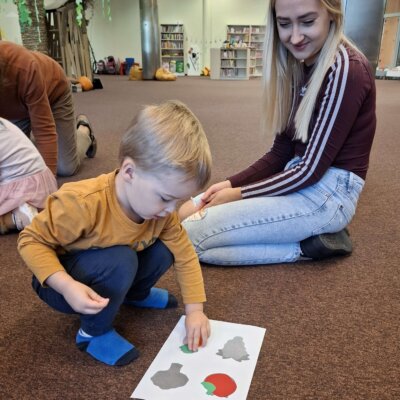 Na fotografii chłopiec pochyla się nad kartką, na której umieszcza wycięte elementy. Obok na wykładzinie siedzi jego opiekun. W tle widać regały z książkami i dekoracje. Kliknięcie w obrazek spowoduje powiększenie.