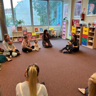 Zdjęcie przedstawia grupę osób siedzących na podłodze w okręgu. W centrum siedzi Pani Bibliotekarka, która czyta książkę. Wokół niej siedzą dzieci wraz z opiekunami. W tle widać duże okna oraz regały z kolorowymi pojemnikami. Na ścianie wiszą plakaty z rysunkami. Kliknięcie powoduje powiększenie zdjęcia.