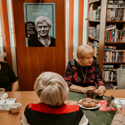 Wnętrze biblioteki. Przy stole widoczne cztery kobiety. Trzy z nich patrzą na moderatorkę spotkania siedzącą poza kadrem. Siedzące przodem czytelniczki uśmiechają się. Kliknięcie w obrazek spowoduje powiększenie obrazu.