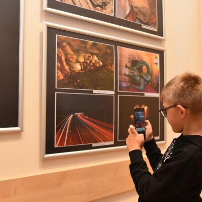 W pomieszczeniu znajduje się wiele osób, które rozmawiają ze sobą i oglądają fotografie wiszące na ścianach. Ludzie są różnie ubrani, niektórzy mają na sobie kolorowe ubrania, a inni bardziej stonowane. W tle widać stół, na którym znajdują się filiżanki i talerzyki. Na ścianach wiszą różne fotografie, które przyciągają uwagę uczestników. Kliknięcie w obrazek spowoduje powiększenie.