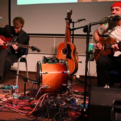 Na zdjęciu znajduje się scena muzyczna z dwoma muzykami. Jeden z nich siedzi po lewej stronie, grając na gitarze elektrycznej. Ma na sobie ciemne ubrania i białe buty. Przed nim znajduje się zestaw perkusyjny z bębnem i różnymi kablami oraz sprzętem elektronicznym. Drugi muzyk siedzi po prawej stronie, grając na gitarze akustycznej. Ma na sobie białą koszulkę i czerwoną czapkę. Obaj muzycy są skoncentrowani na grze na swoich instrumentach. W tle widać ścianę z panelami akustycznymi. Kliknięcie powoduje powiększenie zdjęcia.