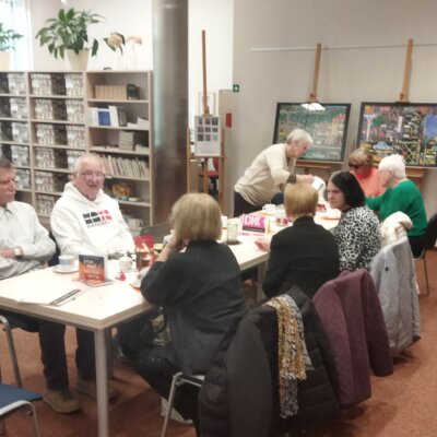 Wnętrze biblioteki, widok stołu po skosie, przy nim siedzą osoby i dyskutują. Jedna stoi i pochyla się nad stołem. Klikniecie powoduje powiększenie zdjęcia.