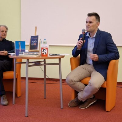 Przy stoliku na pomarańczowych fotelach siedzi autor, Jan M. Piskorski i mówiący do mikrofonu dr Grzegorz Urbanek. Kliknięcie powoduje powiększenie zdjęcia.