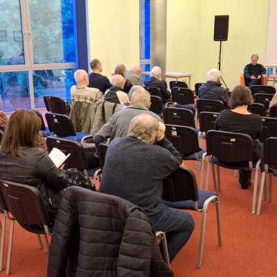 Publiczność zgromadzona w sali. W tle autor i prowadzący spotkanie. Kliknięcie powoduje powiększenie zdjęcia.