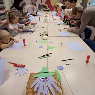 Na zdjęciu widać grupę dzieci i dorosłych siedzących przy długim stole, zajmujących się pracami plastycznymi. Na stole leżą różne materiały do rękodzieła, takie jak kolorowy papier, kleje w sztyfcie, nożyczki i pisaki. Dzieci i dorośli są zaangażowani w wycinanie i sklejanie elementów. Na pierwszym planie, na stole, znajduje się koszyk z położoną na nim papierową postacią przypominającą czarownicę z kapeluszem. W tle na ścianie widoczny jest kolorowy mural z kwiatami i chmurami.