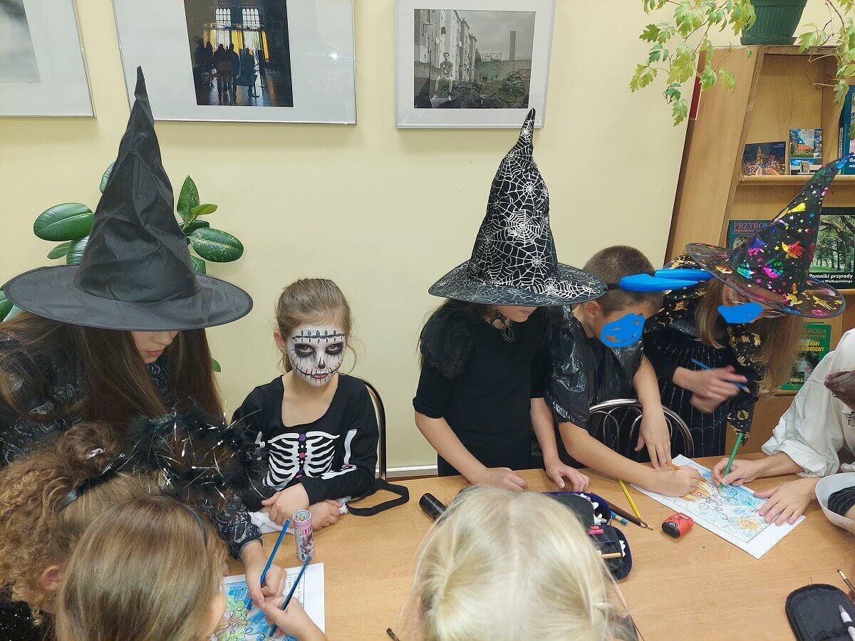 Przy stole dzieci w przebraniach halloweenowych z przewagą koloru czarnego. Na stole kredki i 2 kartki na których dzieci malują wspólną pracę. W tle jasno żółta ściana z fotografiami i zielone kwiaty.