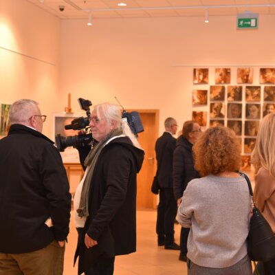 Na zdjęciu znajduje się grupa osób w galerii sztuki. W tle widać ścianę z wieloma obrazami lub fotografiami, które są rozmieszczone w siatce. Osoby na zdjęciu są zwrócone w stronę dzieł sztuki, a jedna z nich trzyma kamerę. Wnętrze galerii jest dobrze oświetlone, a ściany mają jasny kolor. Kliknięcie w obrazek spowoduje powiększenie.