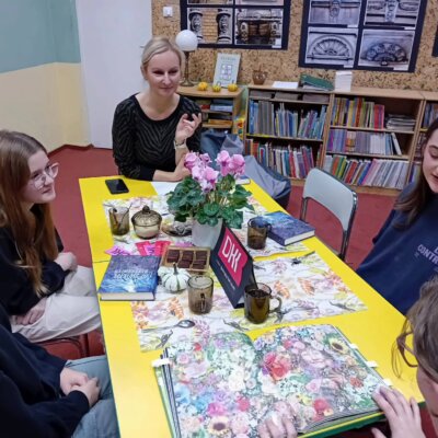 Klubowiczki razem z bibliotekarką siedzą przy stole. Jedna z dziewczynek opisuje kolorową ilustrację, która przed nią leży. Kliknięcie powoduje powiększenie zdjęcia.