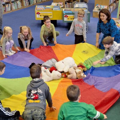 Dzieci razem z bibliotekarką kucają dookoła kolorowej chusty animacyjnej. Na środku chusty leżą misie maskotki. Kliknięcie powoduje powiększenie zdjęcia.