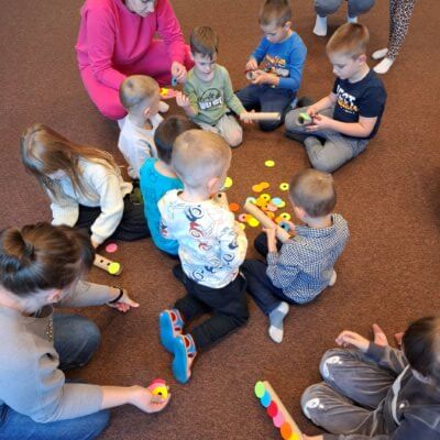 Na fotografii można zauważyć grupę dzieci i dorosłych siedzących na podłodze w pomieszczeniu. Wszyscy skupieni są na wykonywanym zadaniu. Dzieci przy pomocy opiekunów doczepiają kolorowe kółeczka na rzepach do tubek. Kliknięcie w obrazek spowoduje powiększenie.