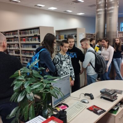 Grupa uczniów zebranych wokół lady bibliotecznej. Słuchają opowieści bibliotekarza. Kliknięcie w obrazek spowoduje powiększenie.