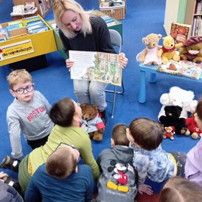 Bibliotekarka pokazuje obrazek w książce o misiu Paddingtonie, a dzieci go oglądają. Kliknięcie powoduje powiększenie zdjęcia.