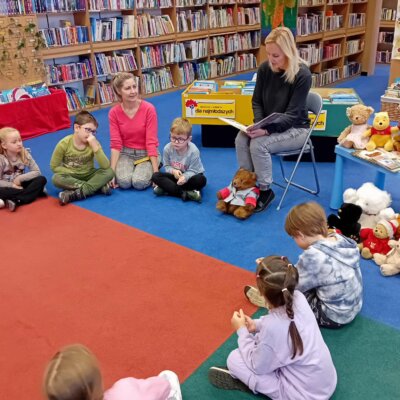 Dzieci siedzą w kółku na dywanie, a bibliotekarka czyta im historię misia Paddingtona. Kliknięcie powoduje powiększenie zdjęcia.