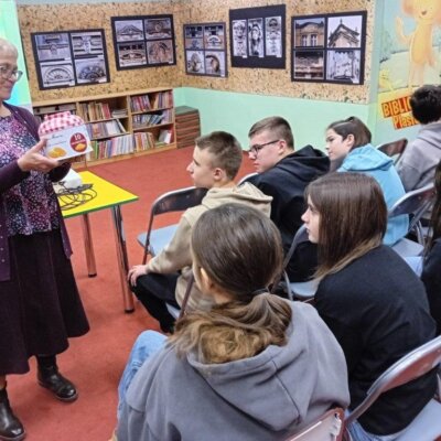 Po prawej stronie uczniowie siedzą na krzesłach. Po lewej stronie prowadząca pokazuje pudełko z ciastkami. Kliknięcie w obrazek spowoduje powiększenie.