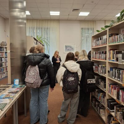 Zdjęcie przedstawia jasne pomieszczenie Oddziału Integracji i Aktywności. Bibliotekarka mówi do młodzieży, która słuch zwrócona w jej kierunku. Po prawej stronie znajdują się regały ze zbiorami bibliotecznymi – płytami CD. Po lewej stronie widoczny jest metalowy filar i stół z prezentacją książek dużym drukiem. Na podłodze pomieszczenia znajduje się jasnobrązowa wykładzina. Za bibliotekarką, na beżowej ścianie znajduje się utrzymany w jasnej tonacji obraz z motywem roślinnym. W oknach wiszą żaluzje pionowe w jasnym kolorze. Widoczny jest fragment sufitu z białych kasetonów. Kliknięcie w obrazek spowoduje powiększenie.