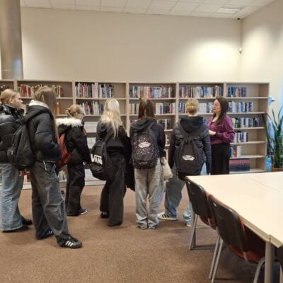 Na zdjęciu znajduje się grupa młodzieży zwrócona w kierunku pracownicy biblioteki opowiadającej o zbiorach Wypożyczalni Głównej. Na wprost za młodzieżą znajdują się regały z książkami (literatura młodzieżowa). Po prawej stronie widać stół w jasnym kolorze i dwa czerwone krzesła i stojąca na ziemi, w donicy, sporych rozmiarów roślina . Za regałami po lewej stronie widoczny jest metalowy filar, ściany pomieszczenia w jasnym, beżowym kolorze i fragment białego sufitu z kasetonów. Na podłodze znajduje się jasnobrązowa wykładzina. Kliknięcie w obrazek spowoduje powiększenie.