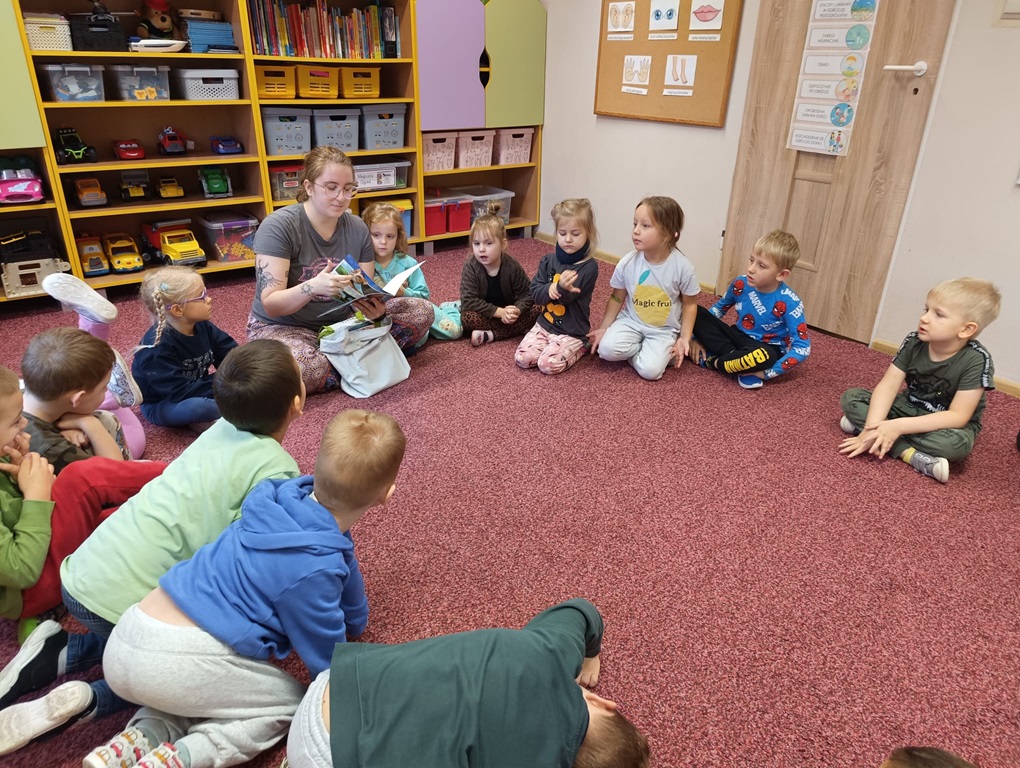 Czternastka dzieci siedząca w kole na czerwonawym dywanie razem z bibliotekarką czytającą książkę na tle dziecięcej meblościanki z zabawkami w przedszkolu.