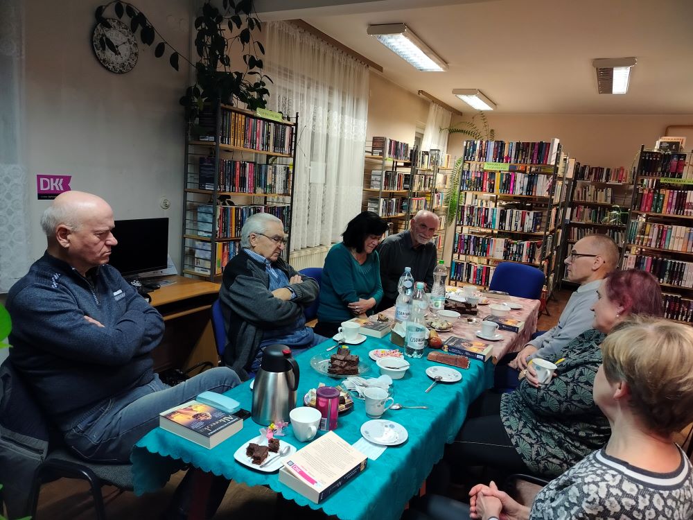 Siedmioro uczestników DKK siedzi przy stole: czterech mężczyzn i trzy kobiety. Na stole są dwa obrusy: zielony i beżowy. Na stole leżą egzemplarze książki „Aktoreczka” Barbary Wysoczańskiej, białe filiżanki i talerzyki, kawa, herbata, słodycze, dzbanek metalowy, ciasto czekoladowe, trzy wody mineralne. W tle są regały z książkami, okna z firankami.