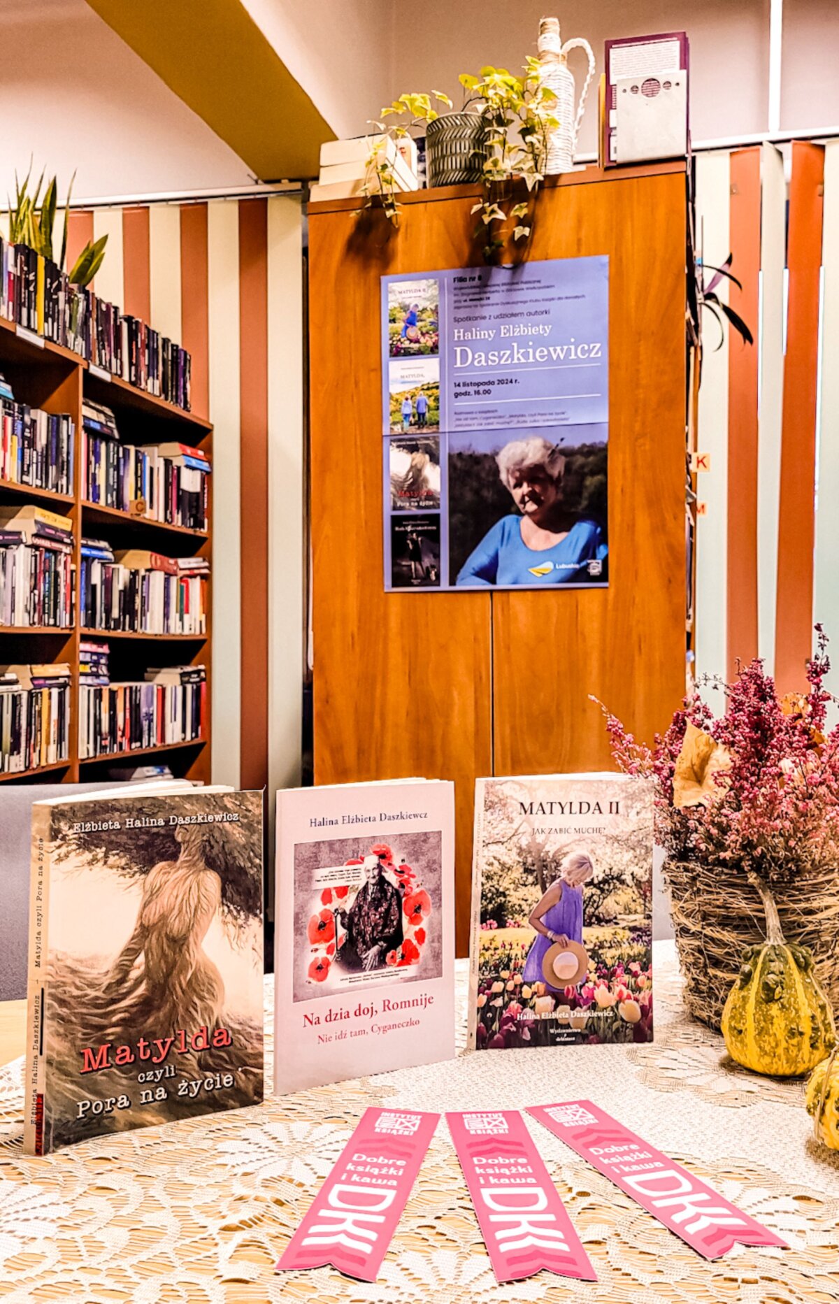 tytułowe – Wnętrze biblioteki na ul. Matejki 38. Na regale plakat zapraszający na spotkanie z Haliną Elżbietą Daszkiewicz – gorzowską powieściopisarką. Na stole trzy książki napisane przez autorkę oraz zakładki promujące Dyskusyjne Kluby Książki.