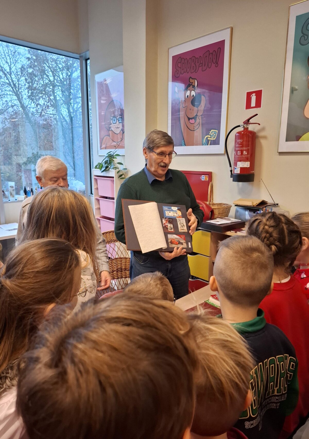 Na zdjęciu znajduje się grupa dzieci stojących wokół dwóch mężczyzn. Jeden z nich trzyma w rękach klaser na znaczki i pokazuje je dzieciom. W tle widać duże okno, przez które można dostrzec drzewa na zewnątrz. Na ścianie za mężczyzną wiszą dwa plakaty z postacią Scooby-Doo. Obok plakatów znajduje się gaśnica. W pomieszczeniu jest także kilka półek z różnymi przedmiotami.