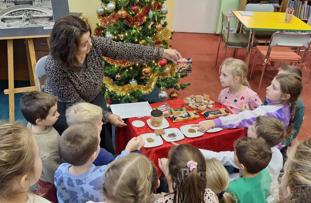 Na czerwonym stoliku na talerzykach leżą różne przyprawy oraz pierniki. Bibliotekarka trzyma torebeczkę gałki muszkatołowej i opowiada, a dzieci zgromadzone dookoła uważnie słuchają.