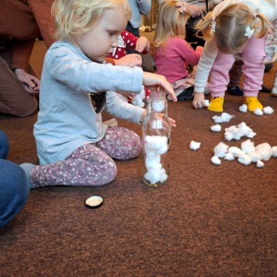 Na zdjęciu znajduje się grupa małych dzieci bawiących się na podłodze. Na pierwszym planie po lewej stronie jest dziewczynka, która wkłada kawałki waty do szklanej butelki. /obok niej na wykładzinie leży zakrętka od butelki. Po prawej stronie inna dziewczynka pochyla się, zbierając kawałki waty z podłogi. W tle widać inne dzieci i dorosłych, którzy obserwują lub pomagają dzieciom w zabawie. Cała scena ma miejsce na brązowej wykładzinie. Kliknięcie w obrazek spowoduje powiększenie.