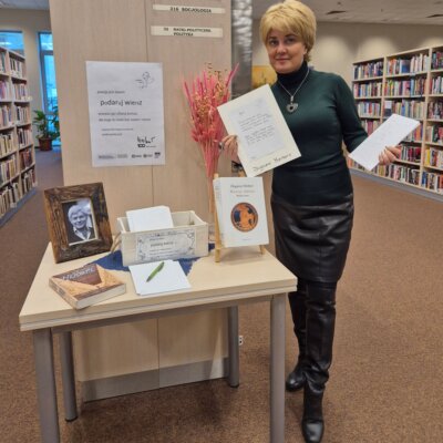 Fragment pomieszczenia Wypożyczalni Głównej biblioteki, w tle widać regały z książkami i wysokie duże okna z widokiem na Park Róż, przy jednym z regałów stoi stolik, a na nim zdjęcie poety Zbigniewa Herberta w ozdobnej ramce oraz koszyczek z papeterią i dwie książki z wierszami poety, w tym jedną na małej sztaludze, obok stolika stoi koordynatorka akcji "Podaruj wiersz" trzymająca w dłoniach przepisane przez czytelników wiersze, koordynatorka ubrana jest w zielony golf i czarną spódnicę. Kliknięcie powoduje powiększenie zdjęcia.