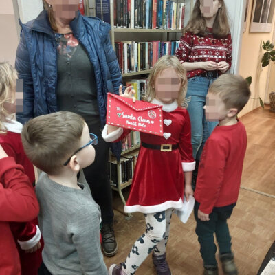 Grupka dzieci i dwie osoby dorosłe. Jedno z dzieci trzyma czerwoną kopertę z napisem Santa Claus. W tle regał z książkami.