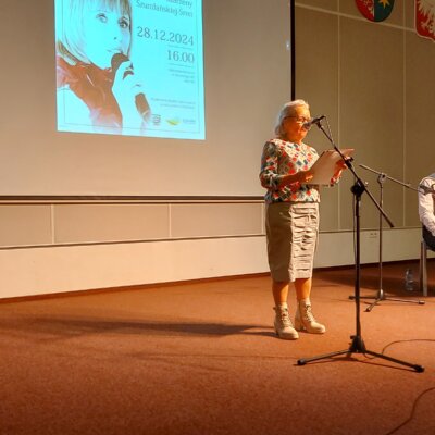 Sala audytoryjna biblioteki, na pierwszym planie za mikrofonem na statywie stoi poetka w kolorowej bluzce i jasnej spódnicy, czyta wiersz, obok na krześle za pulpitem na nuty siedzi akompaniator z akordeonem, za nimi fragment plakatu z koncertu i trzy godła (województwa, kraju i miasta). Kliknięcie powoduje powiększenie zdjęcia.