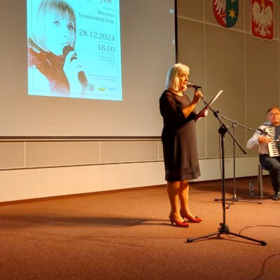 Sala audytoryjna biblioteki, na pierwszym planie za mikrofonem na statywie stoi poetka ubrana w czarną sukienkę i czerwone buty, czyta wiersze, po prawej stronie za pulpitem na nuty siedzi akompaniator z akordeonem, za nimi widać ekran z wyświetlonym fragmentem plakatu z koncertu, obok trzy godła (województwa, kraju i miasta). Kliknięcie powoduje powiększenie zdjęcia.