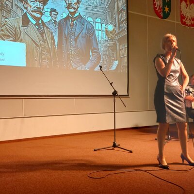 Sala audytoryjna biblioteki, na pierwszym planie stoi wokalistka przebrana w srebrną suknię, wykonuje piosenkę pt. Milord trzymając w lewej dłoni mikrofon, po prawej stronie na krześle za pulpitem na nuty gra akompaniator na akordeonie, za ich plecami widać ekran przedstawiający dwóch panów w eleganckich garniturach z początku XX w., obok wiszą trzy godła (województwa, kraju i miasta). Kliknięcie powoduje powiększenie zdjęcia.