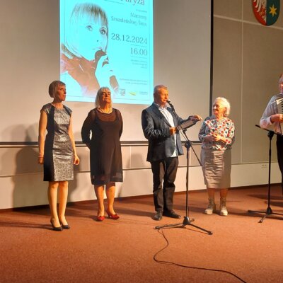 Sala audytoryjna, na pierwszym planie stoją wszyscy wykonawcy koncertu oraz pracownik biblioteki odpowiedzialny za dźwięk i oświetlenie na koncercie, za nimi na ekranie plakat z koncertu i obok trzy godła (województwa, kraju i miasta). Kliknięcie powoduje powiększenie zdjęcia.