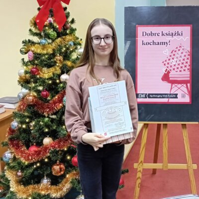 Laureatka konkursu na recenzję pokazuje list gratulacyjny. Po jej lewej stronie stoi ubrana choinka, a po prawej stronie sztaluga z plakatem DKK. Kliknięcie w obrazek spowoduje powiększenie.