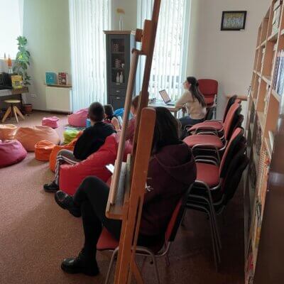 Wnętrze biblioteki, na kolorowych pufach siedzą uczestnicy warsztatów i oglądają slajd wyświetlany na ścianie. Na pierwszym planie duża sztaluga z obrazem. Kliknięcie powoduje powiększenie zdjęcia.