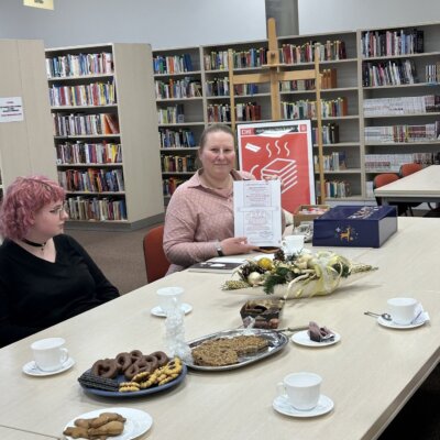 Na zdjęciu laureatka konkursu siedzi przy stole i pokazuje dyplom. Obok niej siedzi jedna z uczestniczek spotkania. W tle widać regały z książkami oraz plakat DKK, który stoi na sztaludze. Kliknięcie powoduje powiększenie zdjęcia.