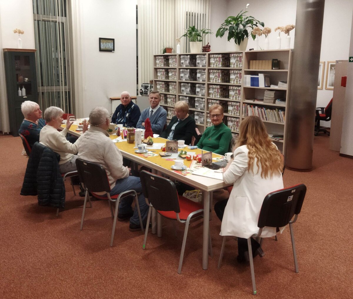 Wnętrze biblioteki, długi stół, nakryty żółtym obrusem, zastawiony filiżankami i poczęstunkiem, przy nim klubowicze dyskutują o książce. W tle regały.