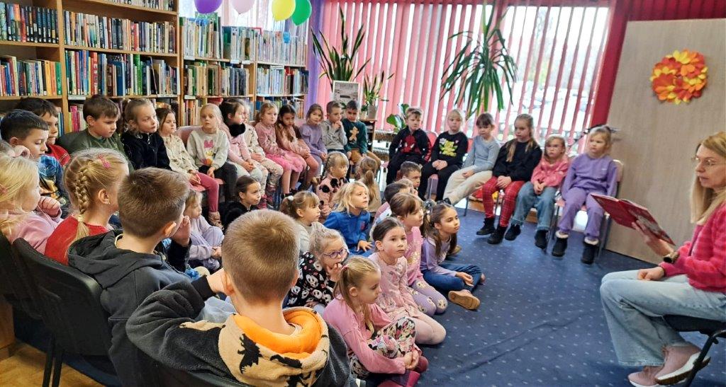 Kobieta czyta grupie dzieci, które siedzą w półokręgu na podłodze. Za nimi regały z książkami.