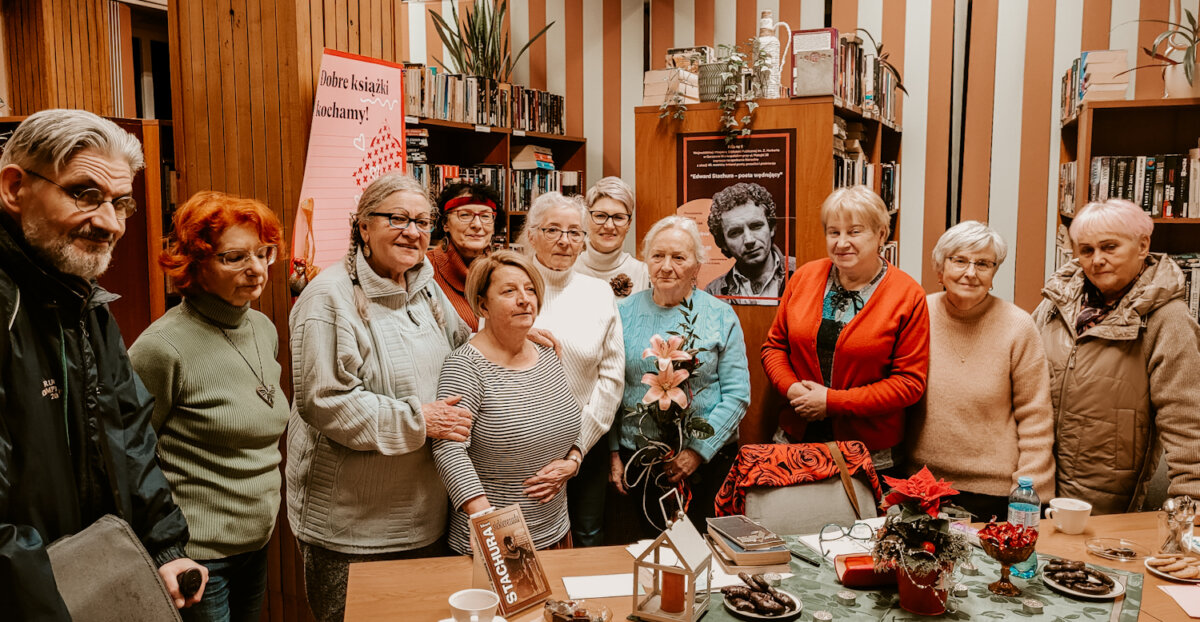 Wnętrze biblioteki. Trwa spotkanie literackie z okazji 45. rocznicy śmierci Edwarda Stachury. Prowadząca Janina Jurgowiak w środku, po bokach stoją uczestnicy spotkania.